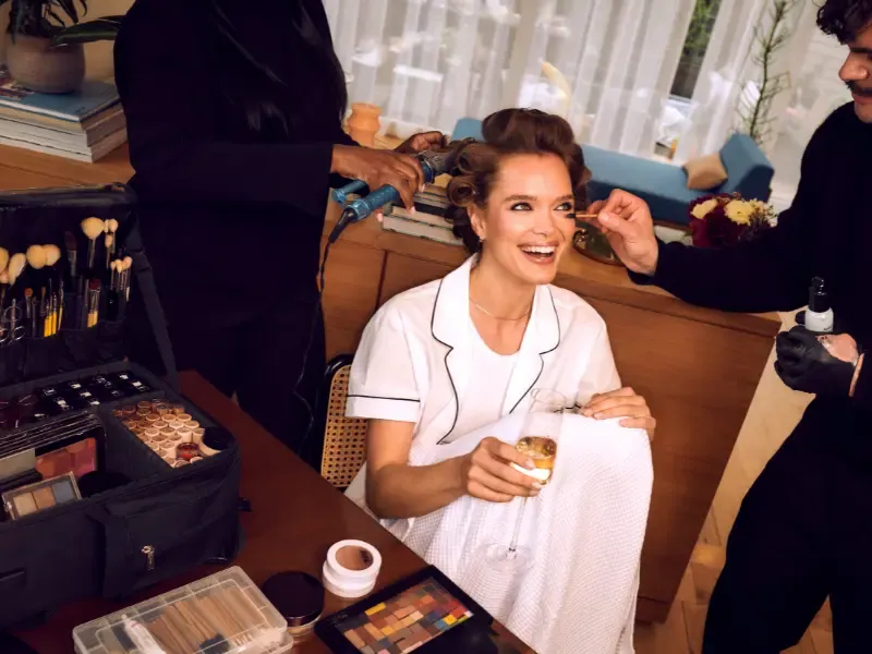 A laughing woman in a white robe enjoys champagne while receiving hair and beauty services from a professional glam team.