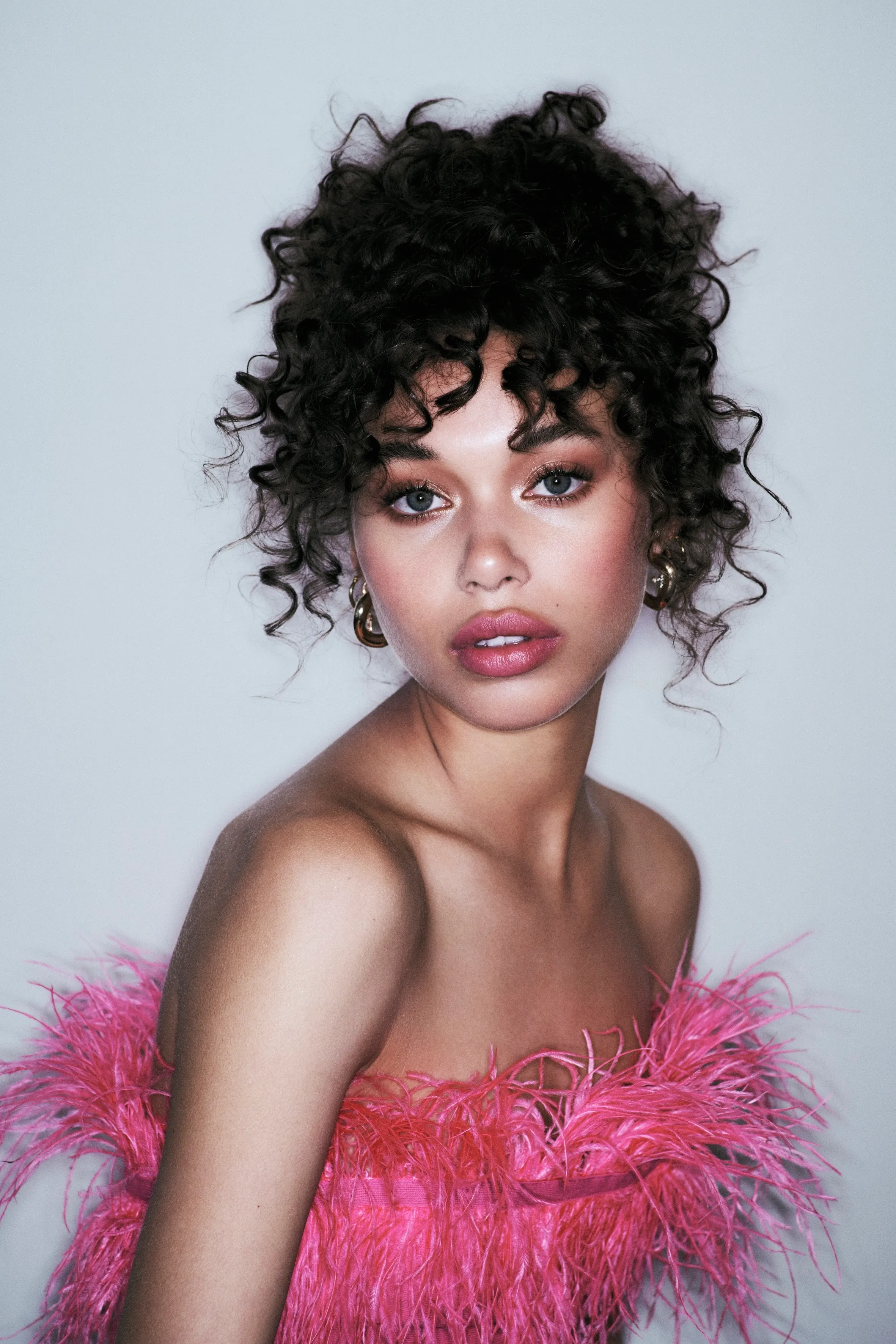 Close-up of a woman in a pink feather top wearing a voluminous curly evening upstyle with gold hoop earrings.