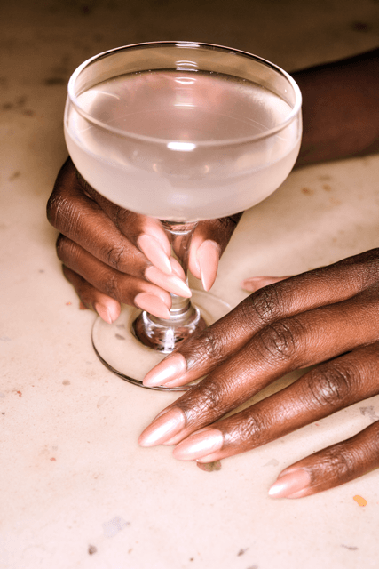 Close-up of elegant hands with long almond-shaped gel manicure in a sheer pink finish, holding a coupe cocktail glass.