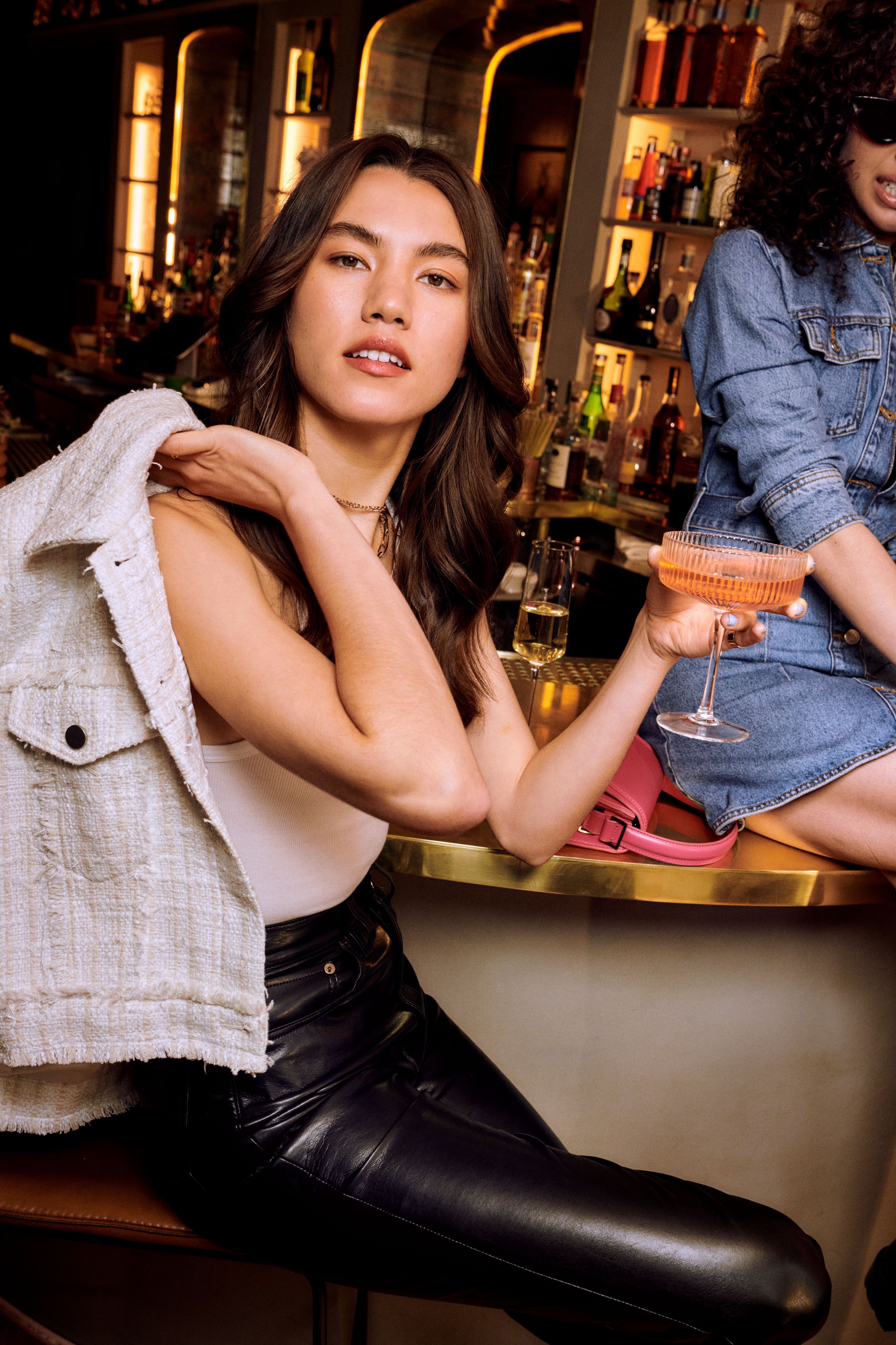 Makeup services bar-ready look brunette woman holding a pink coupe glass while seated at a gold bar counter.