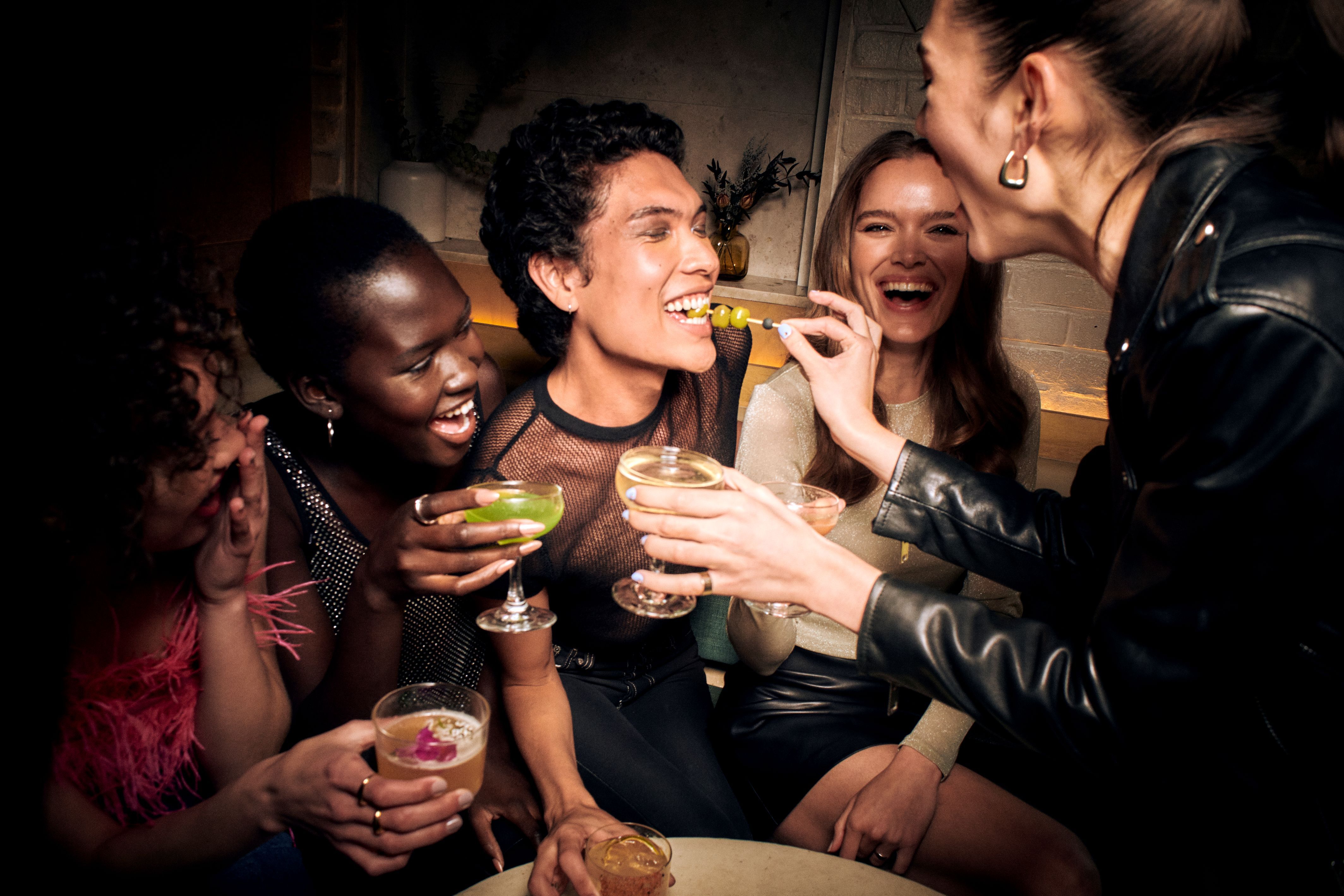 Makeup services group of women laughing and celebrating together holding cocktail glasses at a dimly lit bar.