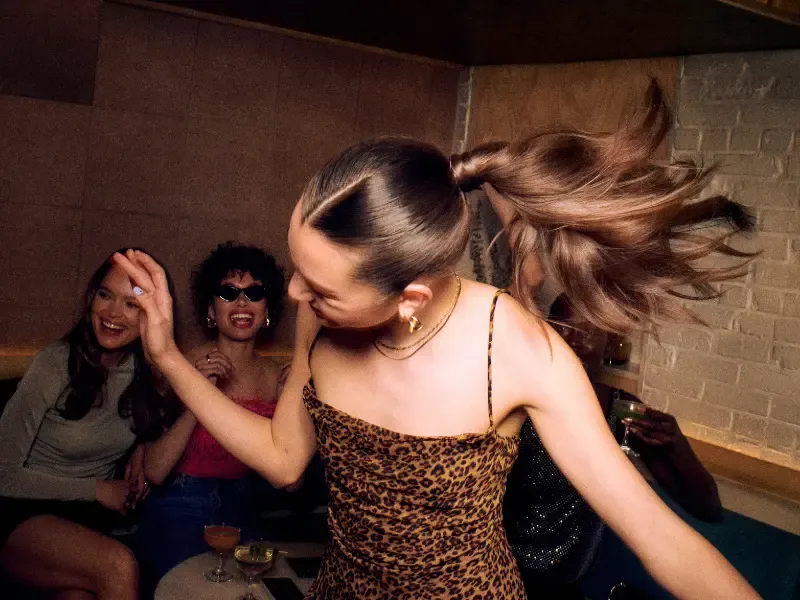 At home hair stylists creating a sleek ponytail for a woman dancing energetically with friends at a lively party