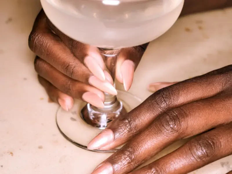 At home manicure service featuring elegant nude almond-shaped nails holding a cocktail glass on a marble surface.