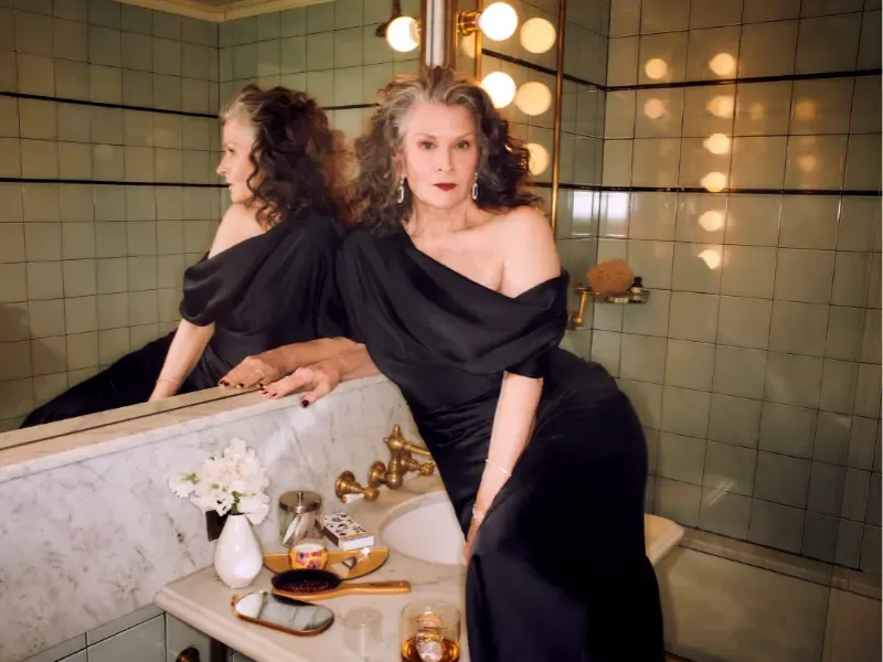 Blowout at home styling shown by a woman with voluminous hair posing confidently in a black gown by a vanity mirror