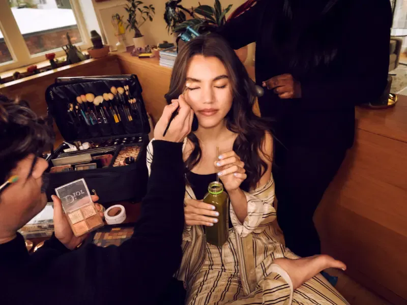Bridal hair and makeup artist perfecting a bride’s look as she sips green juice during her beauty session.