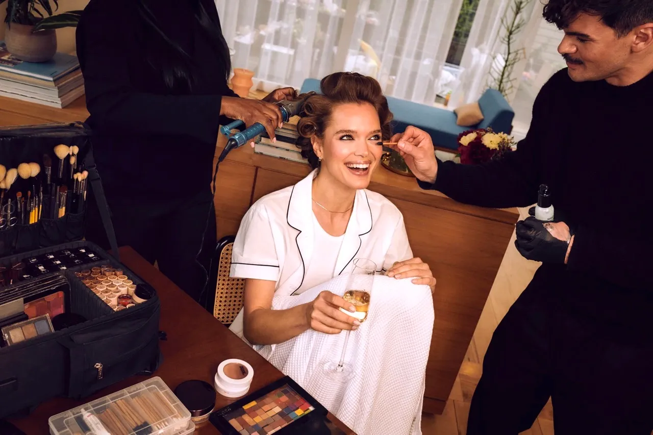 Classic bridal makeup being applied to a smiling bride in pajamas while getting her hair curled