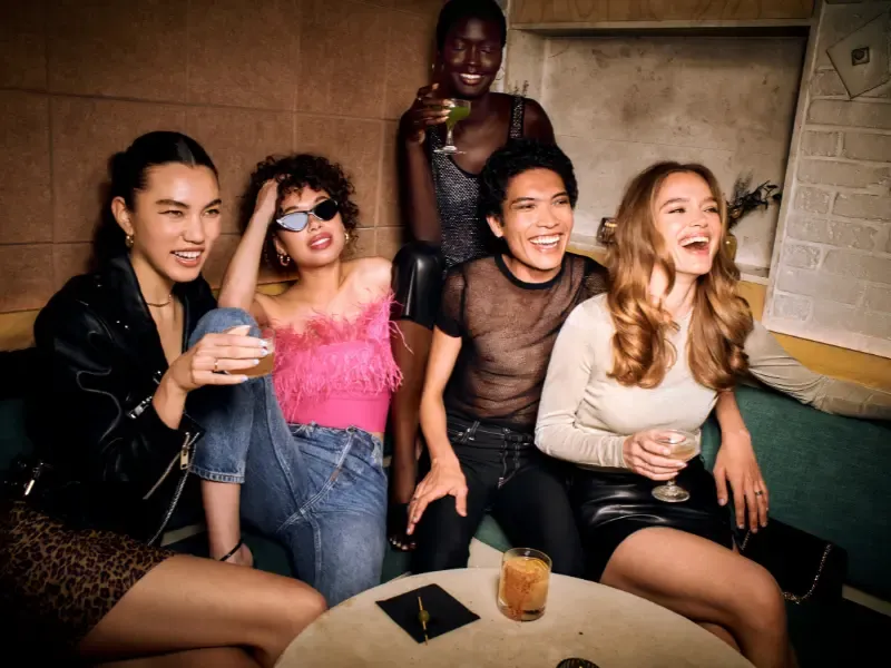Full glam makeup look on a group of friends laughing and enjoying cocktails in a stylish lounge setting.
