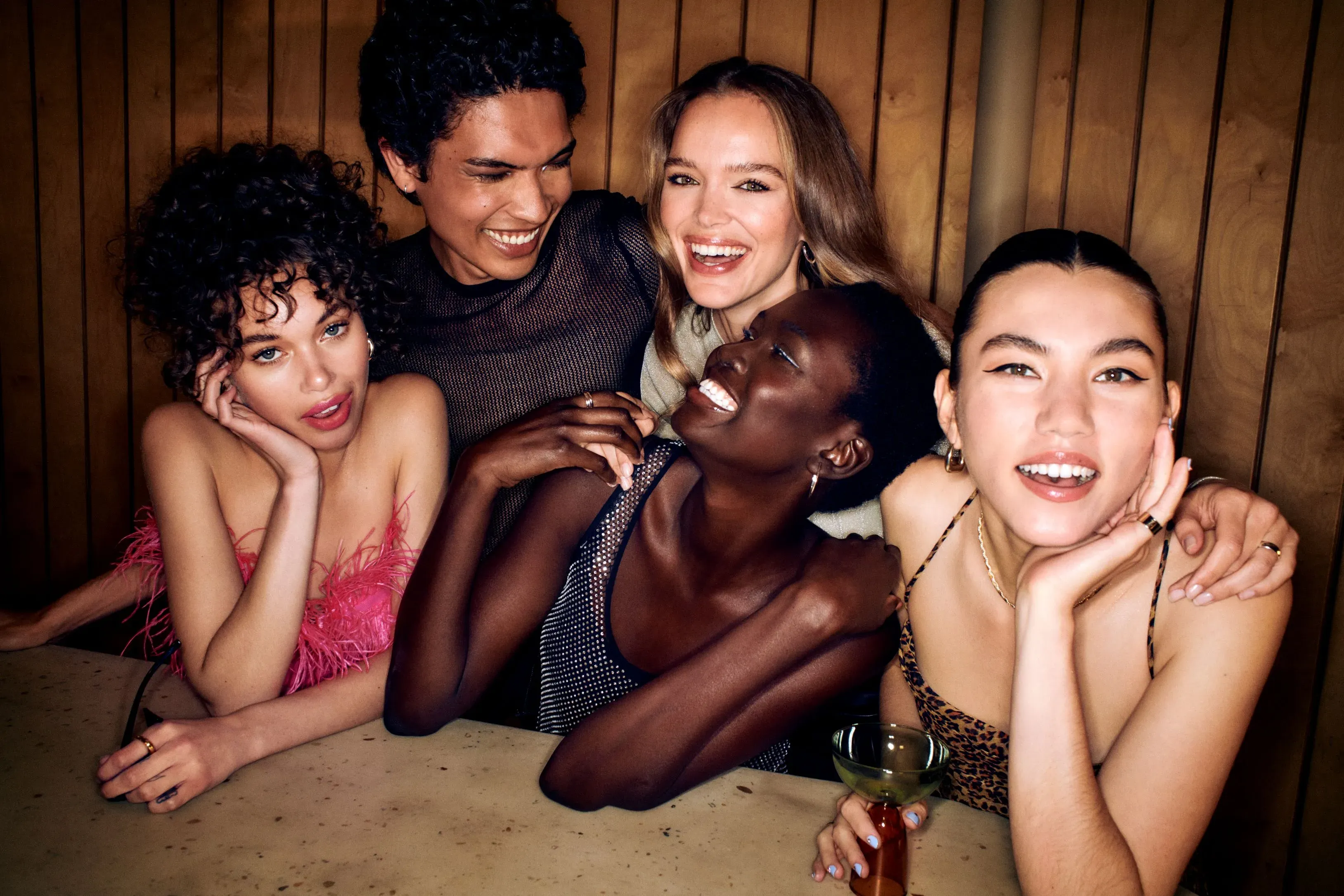 Glam services for a group of five diverse friends laughing and leaning together at a bar booth in a warmly lit venue.