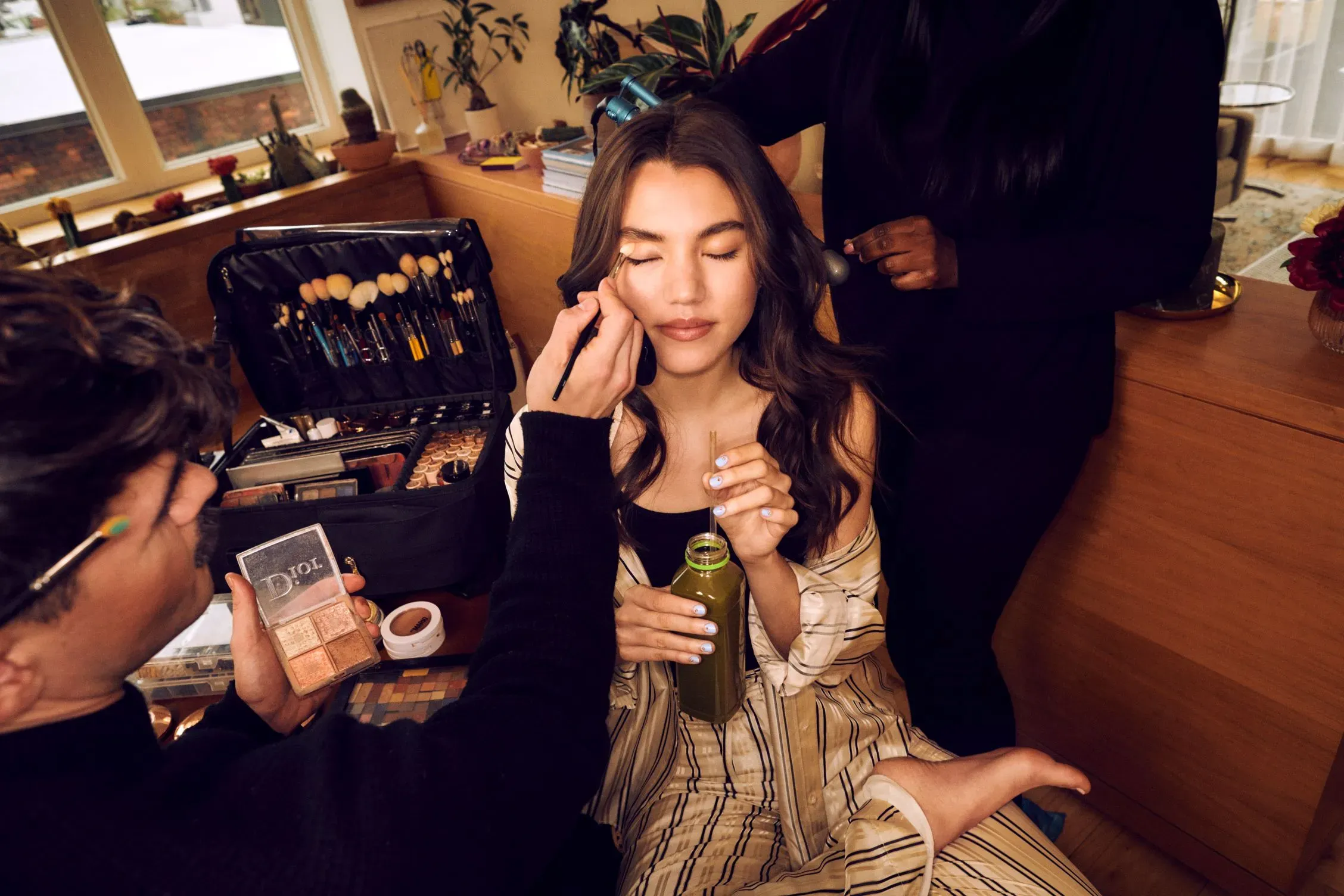 Glam stylists for event apply makeup and style hair as woman relaxes in loungewear sipping green juice.