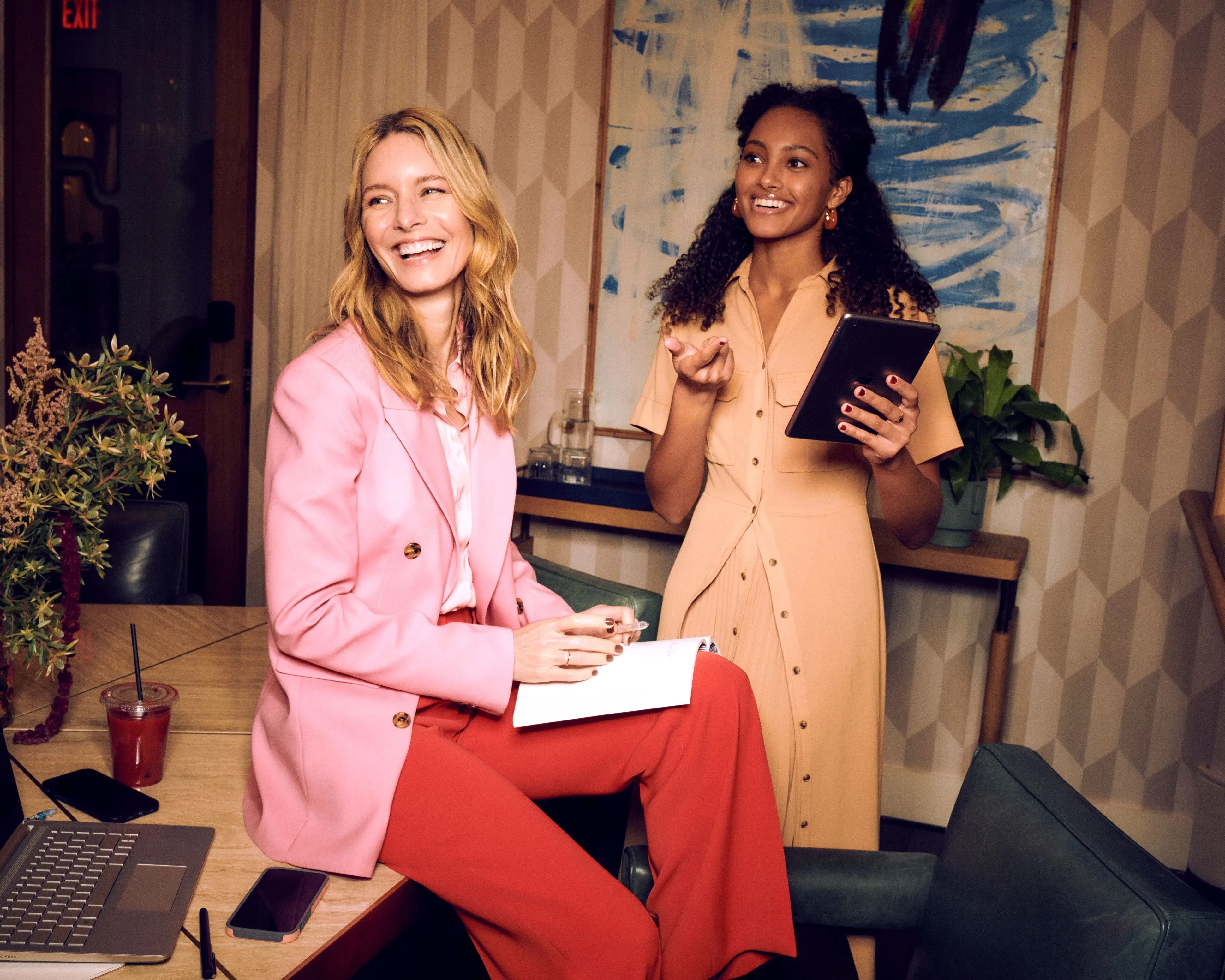 Group glam services with two professionally styled women laughing together in a modern office setting, one in a pink blazer and one in a tan dress holding a tablet.