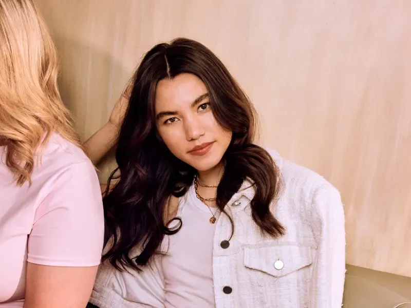 Hair blowout with soft waves on a woman wearing a white jacket and layered necklaces, leaning against a wall.