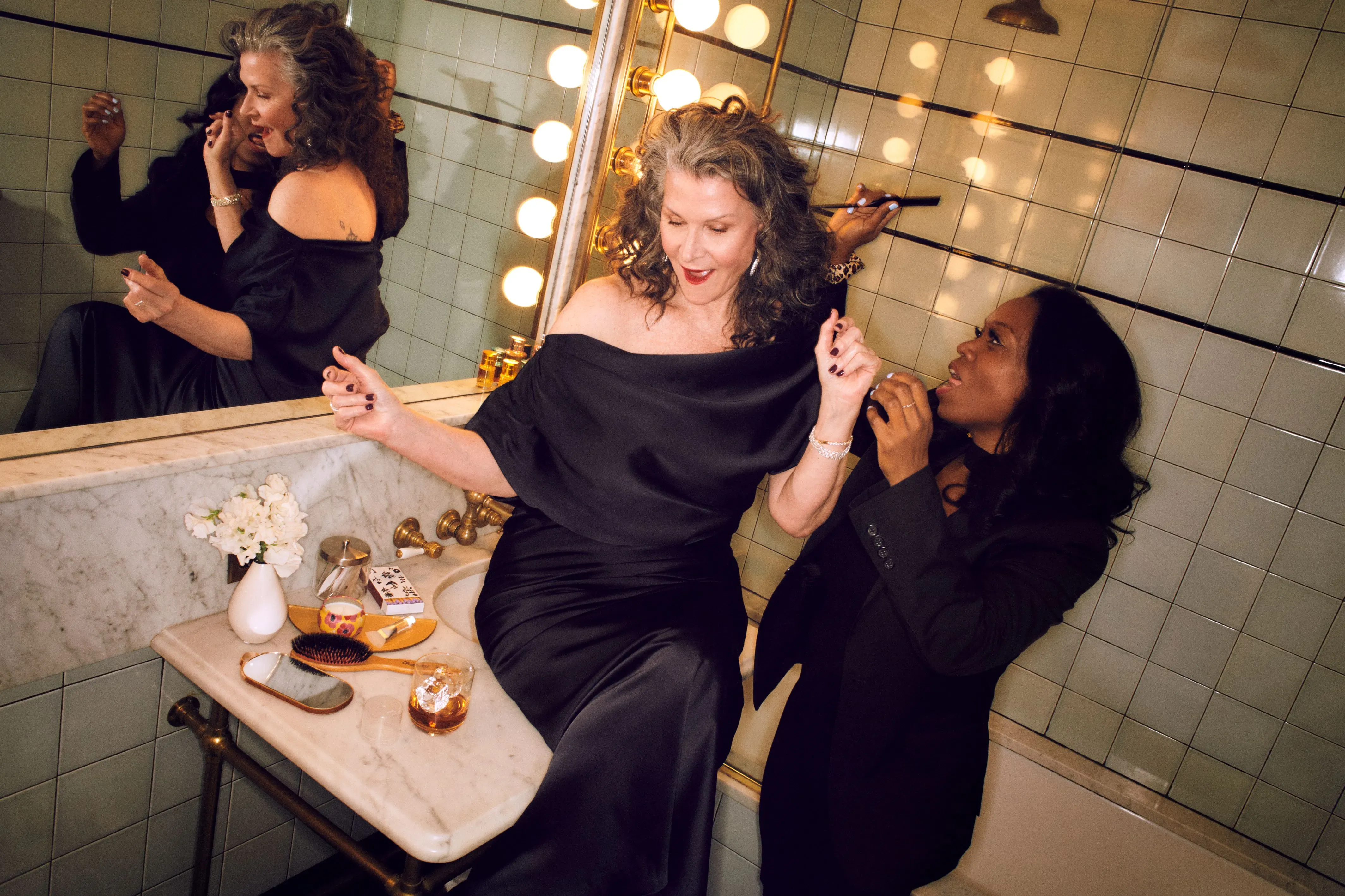 Hair styling services leave a woman in a black silk dress laughing with her stylist in a vanity-lit bathroom before a night out.