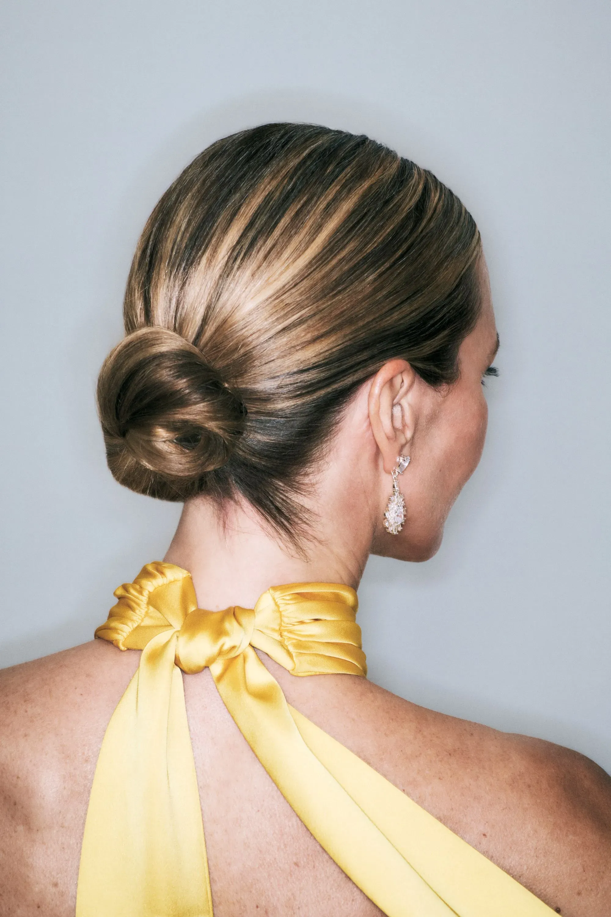 Hairstyles for daily wear showcasing a sleek low bun with highlighted brunette hair, paired with a yellow satin halter dress and crystal drop earrings