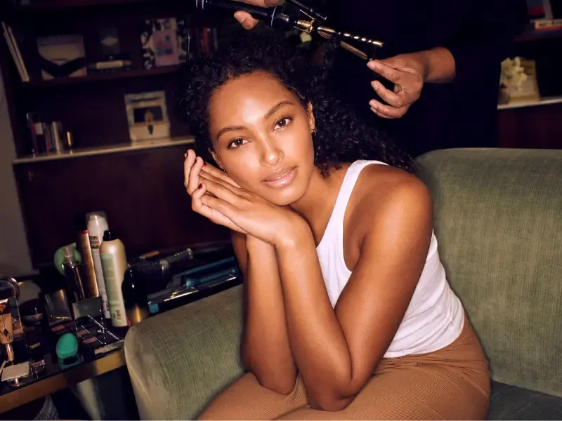 Makeup for a wedding being prepared as a woman with glowing skin and natural makeup sits in a green chair while a stylist curls her hair, surrounded by beauty products on a vanity