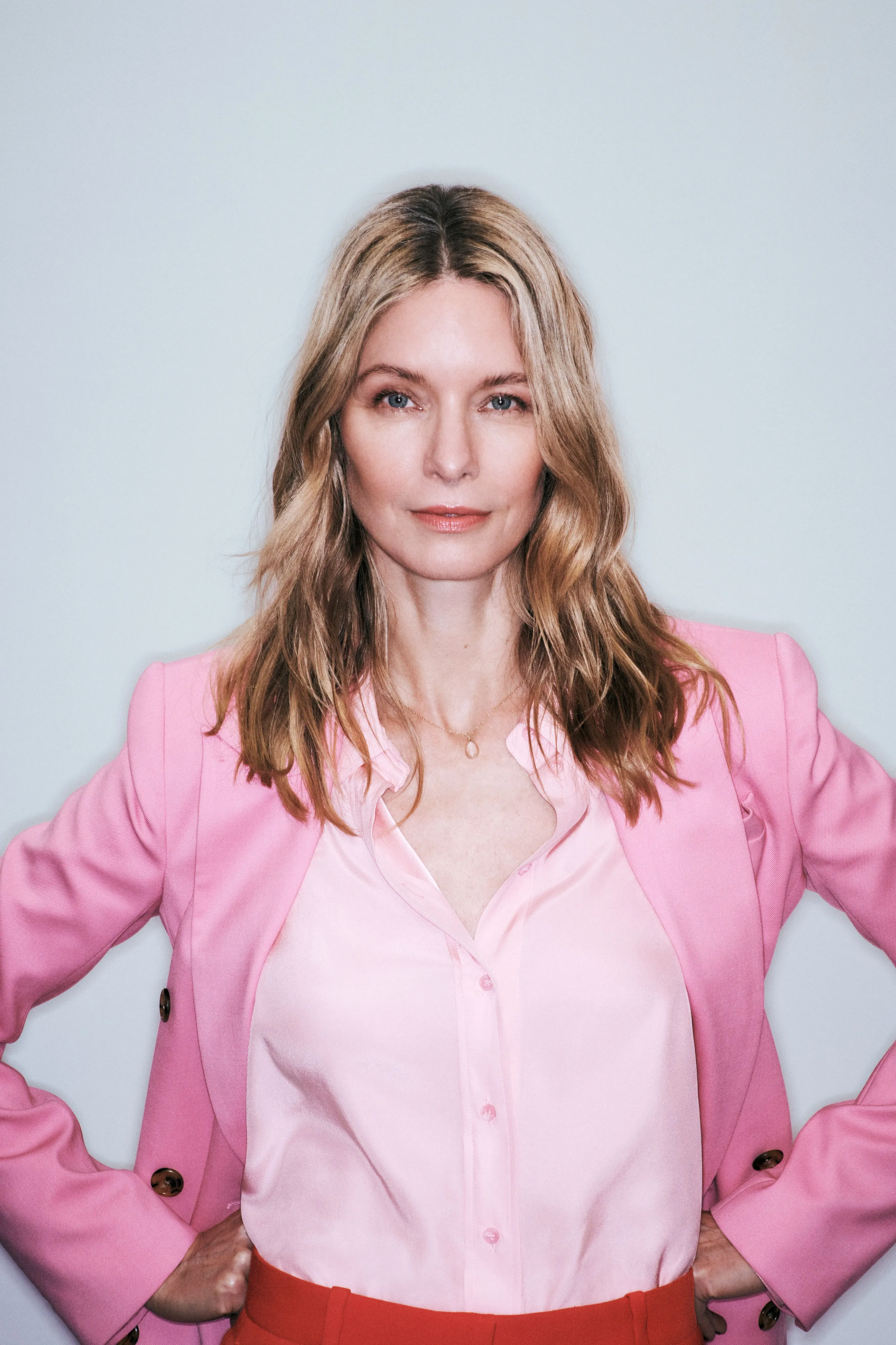 Makeup for corporate event featuring natural glam with soft bronzed skin and wavy blonde hair on a woman in a pink blazer against a light backdrop.