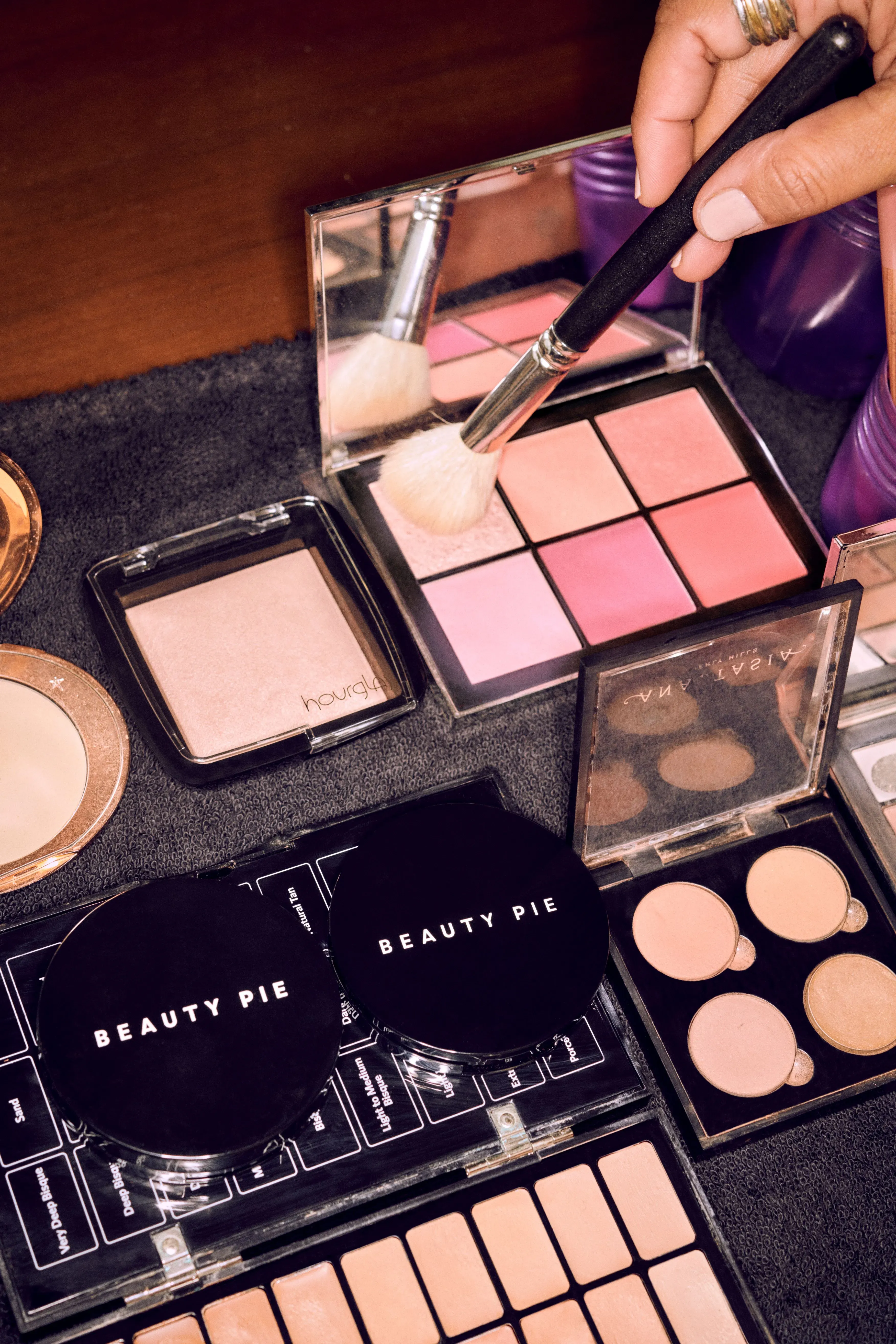 Makeup services flatlay with a brush applying blush from a pink palette surrounded by Beauty Pie compacts, highlighter, and concealer products on a vanity.