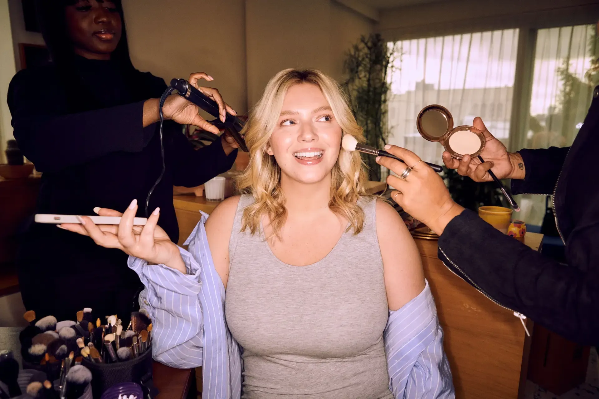 Modern bridal makeup being applied as a woman smiles while getting her hair curled and foundation brushed on.