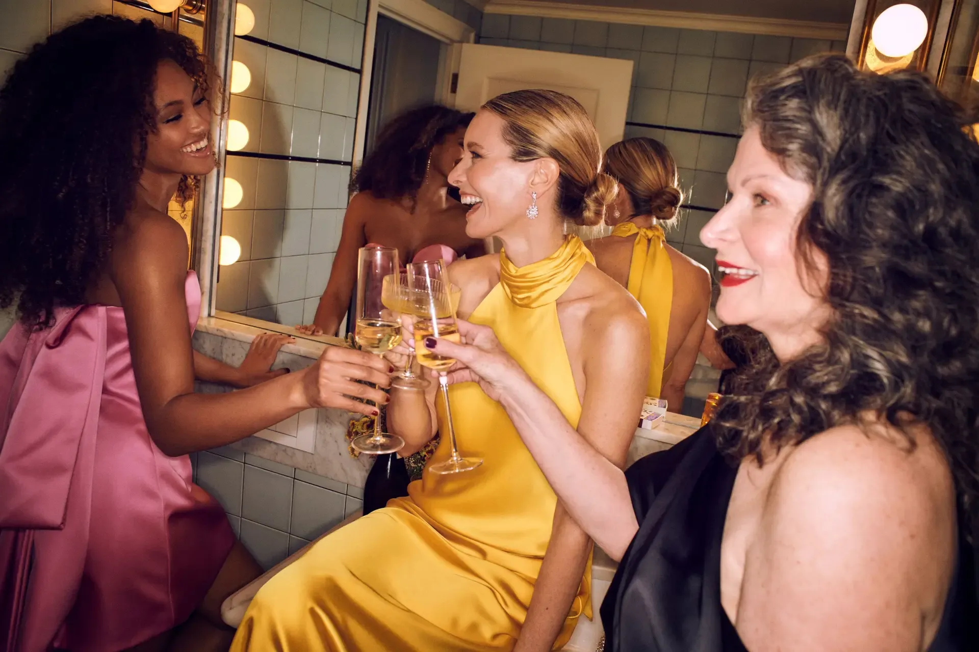 Natural wedding makeup looks are showcased as women toast in a warmly lit vanity room before a celebration.