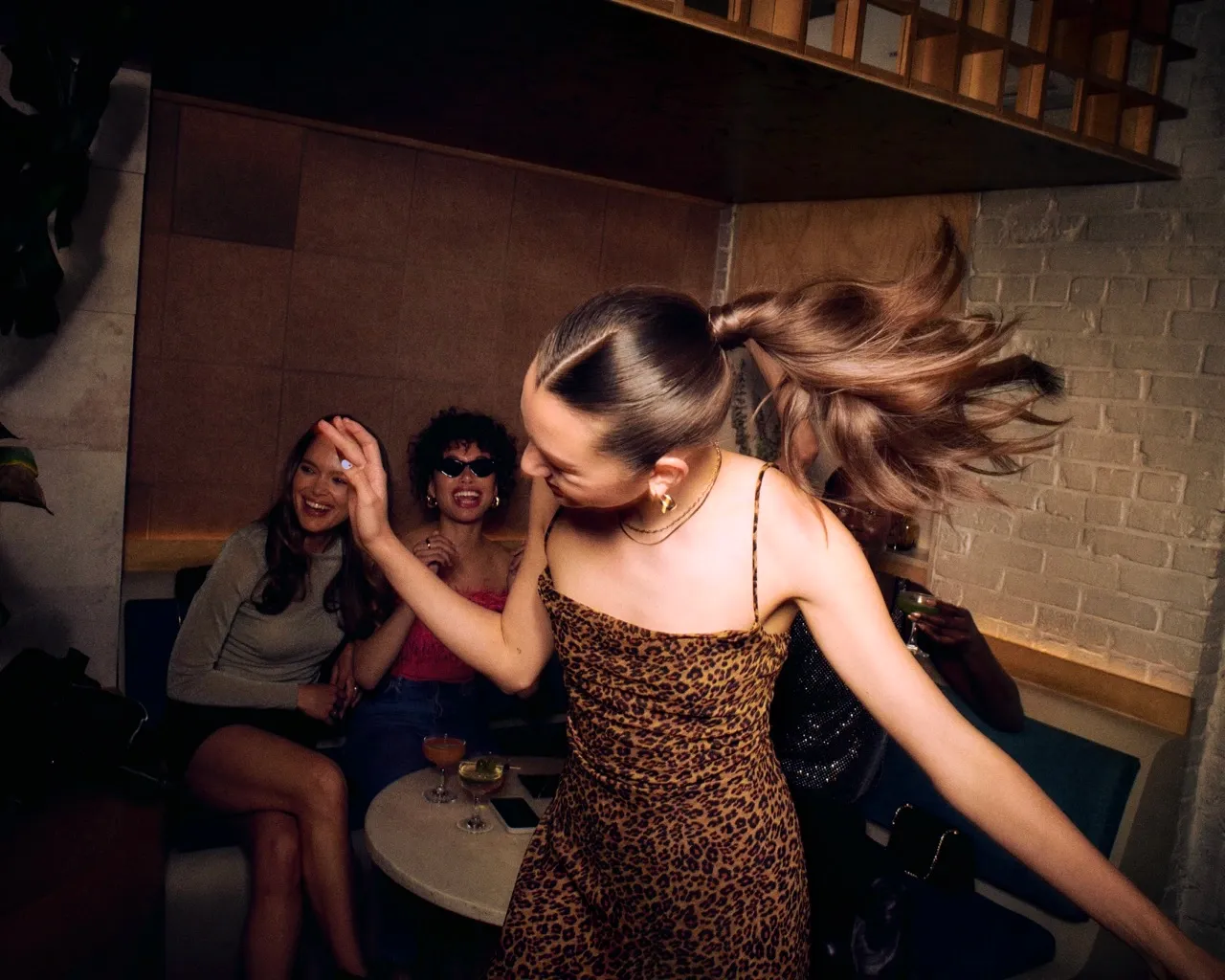 Professional hair service with hairstylists styling a woman's hair while others enjoy drinks at a trendy lounge