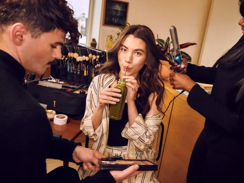 Salon styling at-home while woman sips green juice during hair curling and makeup application.