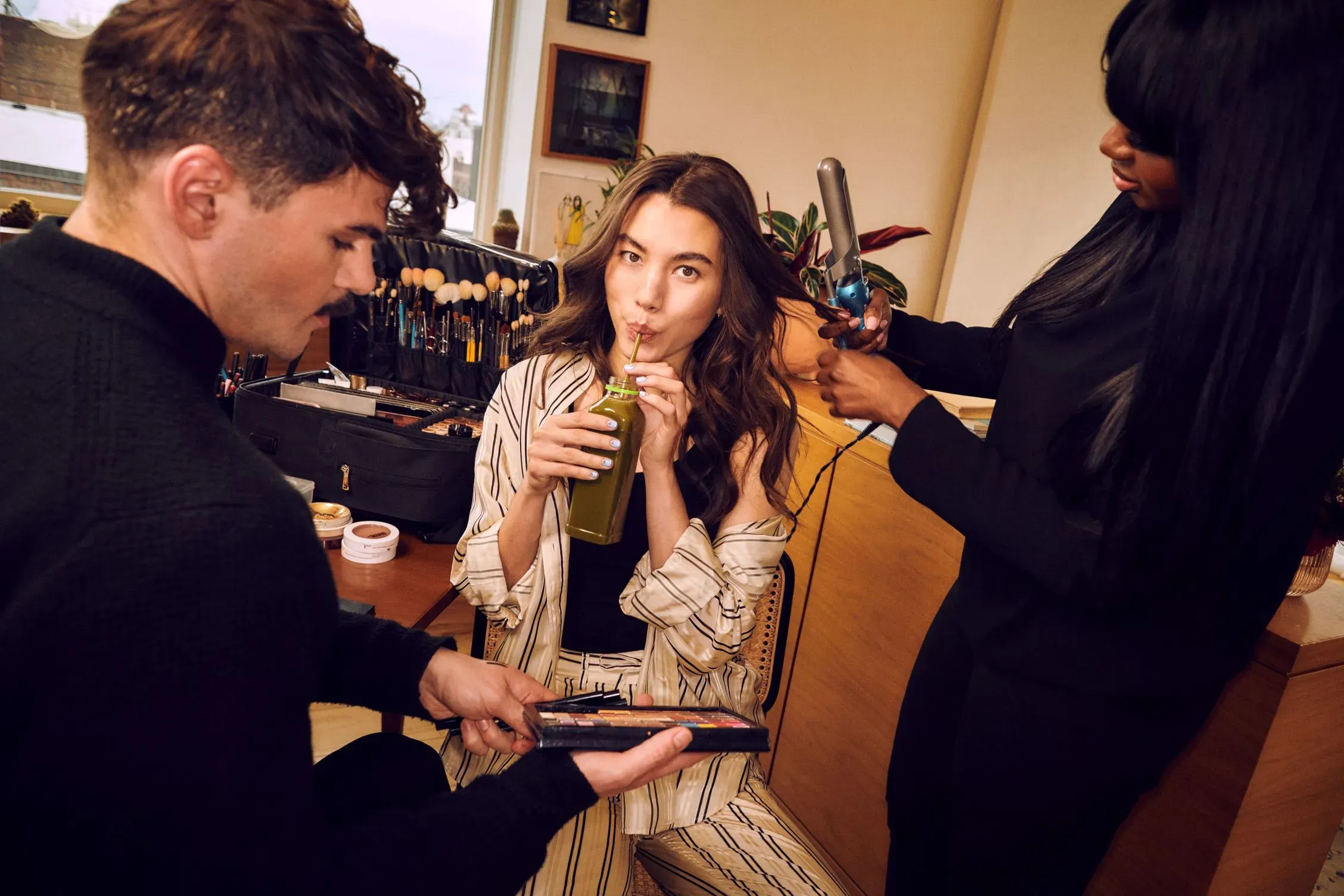 Trending hairstyles being created during a glam session as a woman sips juice and gets her hair styled.